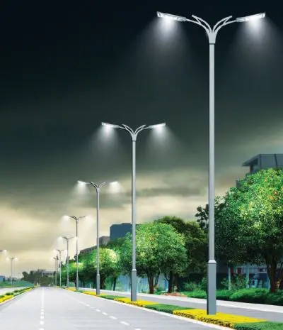 Fila de postes de luz ornamentales de ORBE - Postes Metalicos, con un diseño elegante de doble brazo y luminarias LED que iluminan una avenida moderna al anochecer.